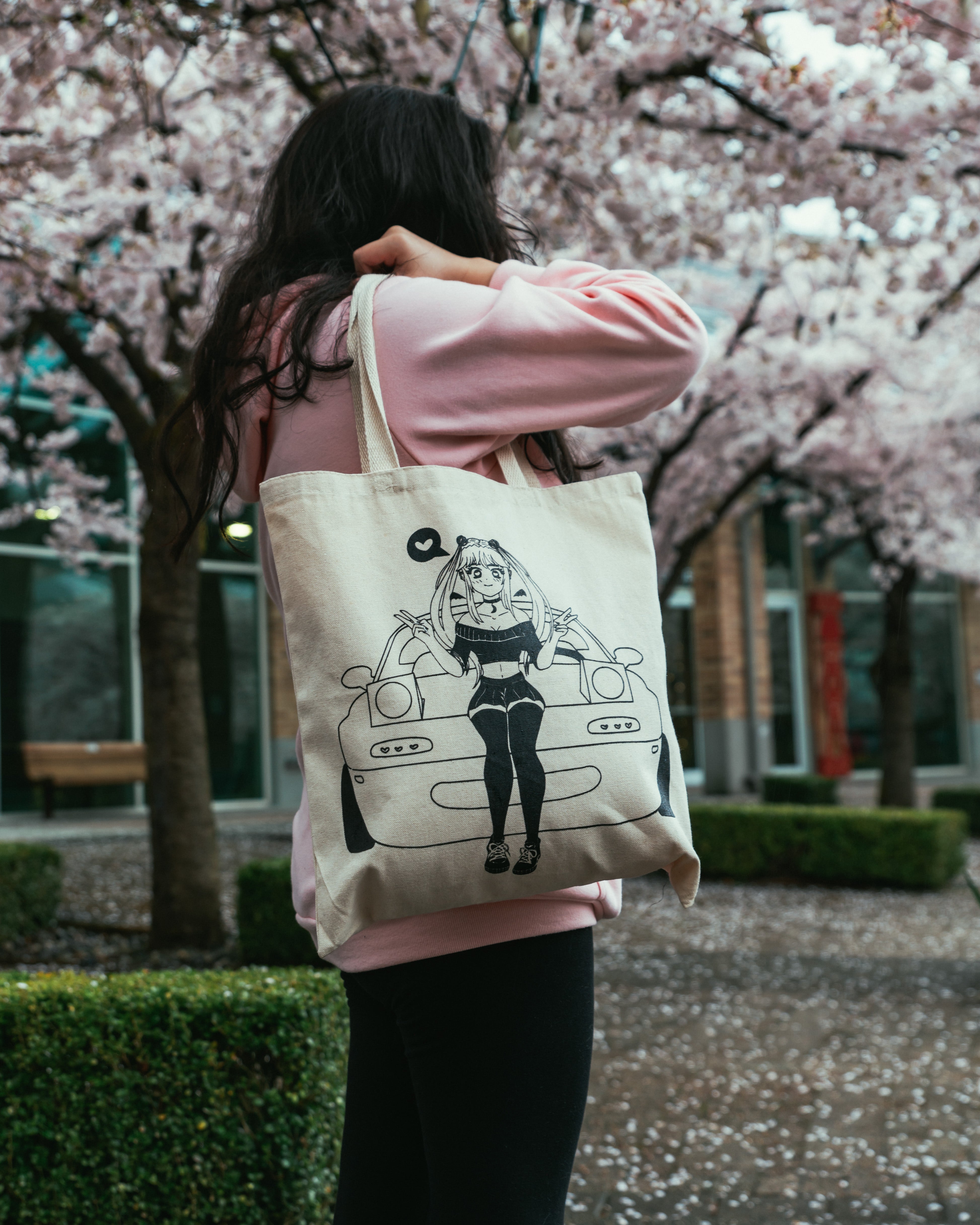 girl wearing a pink hoodie near cherry blossom trees holding a cute tote bag featuring a graphic of an anime girl that looks like sailor moon standing infront of an NA miata